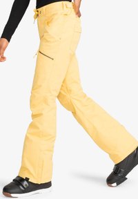 Gelbe Schnee-Hosen mit einem lässigen Schnitt, Cargo-Taschen, Reißverschlüssen an den Seiten und verstellbarem Bund; kombiniert mit schwarzen Schneestiefeln.