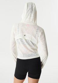 Haut à capuche léger et transparent de couleur blanche avec une découpe dans le dos, une capuche attachée et un petit logo. Assorti à un short noir moulant.