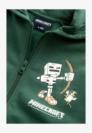 Grön hoodie med ett Minecraft-karaktärsgrafik, med en kubisk figur som håller en båge och en liten vit fågel; inkluderar dragkedja och etikett.