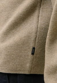 Bežani pleteni materijal s vidljivim šavom i malom crnom etiketom označenom "PREMIUM" blizu ruba, iznad tamnog teksturiranog materijala.