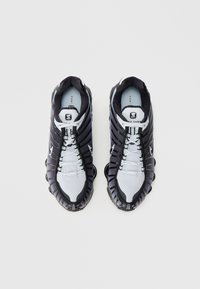 Černo-šedé tenisky Nike Shox s texturovaným síťovaným svrškem, výraznou pružinovou podrážkou a reflexními doplňky; tkaničky jsou bílé.