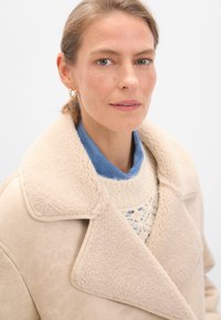 Cappotto in shearling beige con ampio colletto, indossato sopra una camicia blu e un maglione lavorato a maglia con un motivo in pizzo. Orecchini a cerchio dorati aggiungono un tocco di dettaglio.