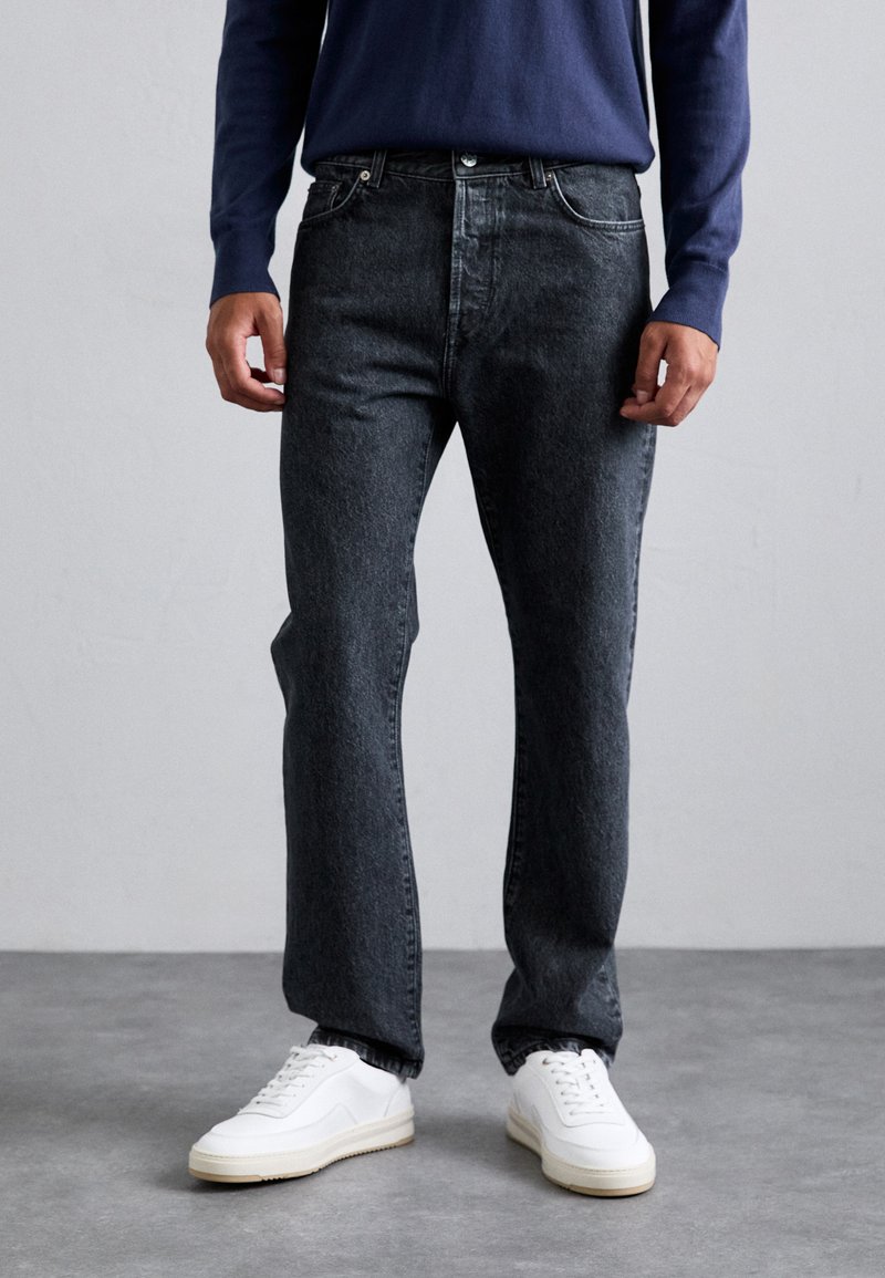 Person wearing black denim jeans, white sneakers, and a navy blue long-sleeve shirt standing on a gray floor and light gray wall background.