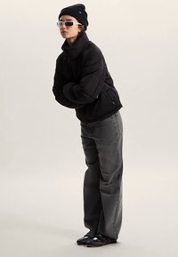 Doudoune noire, jean gris à jambes larges, bonnet noir, lunettes de soleil transparentes, et chaussures noires à brides. Se tient les bras croisés.