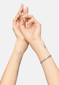 Bracciale d'argento con un design testurizzato, indossato su un polso. Due mani intrecciate, che mostrano tonalità di pelle chiara e tatuaggi floreali su un avambraccio.