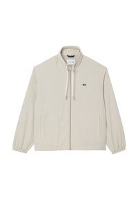 Helle beige Zip-Jacke mit hohem Kragen, elastischen Bündchen, Seitentaschen und grünem Lacoste-Logo auf der linken Brust. Glatte Textur.