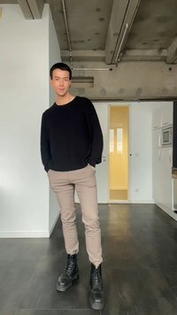 Jonge man in zwarte trui, beige broek en zwarte laarzen staat met handen in zijn zakken in een moderne keuken met betonnen plafond.