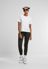 T-shirt en coton blanc, à manches courtes, associé à des leggings noirs. Le mannequin porte des baskets blanches chunky avec des couleurs d'accent visibles et des lunettes de soleil.