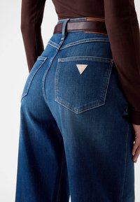 Blauwe denim jeans met een brede pijp, hoge taille en twee achterzakken met een driehoekig merklabel. Een bruin leren riem accentueert de taille.