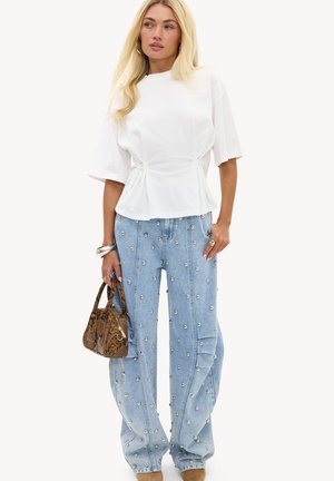 Femme blonde portant un haut blanc cintré à manches courtes, un jean bleu clair avec des clous argentés, tenant un sac à main en cuir de serpent marron.