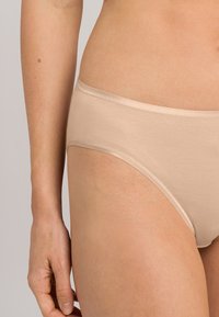 Slip in cotone beige con una texture liscia, vita media e dettagli cuciti lungo i bordi. Aperture delle gambe leggermente curve.