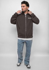 Brown zip-up hoodie with kangaroo pockets, featuring white logo. Paired with light blue distressed jeans and black-and-white sneakers.