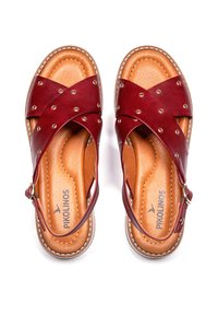Sandalias de cuero rojo con correas cruzadas y tachuelas decorativas de metal. Presenta una plantilla de cuero marrón y una correa ajustable en el tobillo con hebilla.