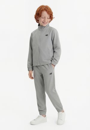 Niño sonriente con cabello rojo que lleva un chándal gris de New Balance y zapatillas blancas, de pie con una mano en el bolsillo sobre un fondo blanco.