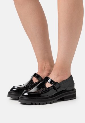 Černé lakované Mary Jane boty s chunky podrážkami, nošené na barevných nohách na jednobarevném bílém pozadí.