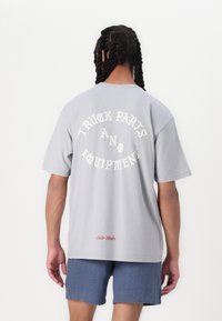 Szary bawełnowy t-shirt z krótkim rękawem, z białym napisem "CZĘŚCI I WYPOSAŻENIE CIĄGNIKÓW" w zakrzywionym wzorze na plecach. Czerwony detal "Auto Works".