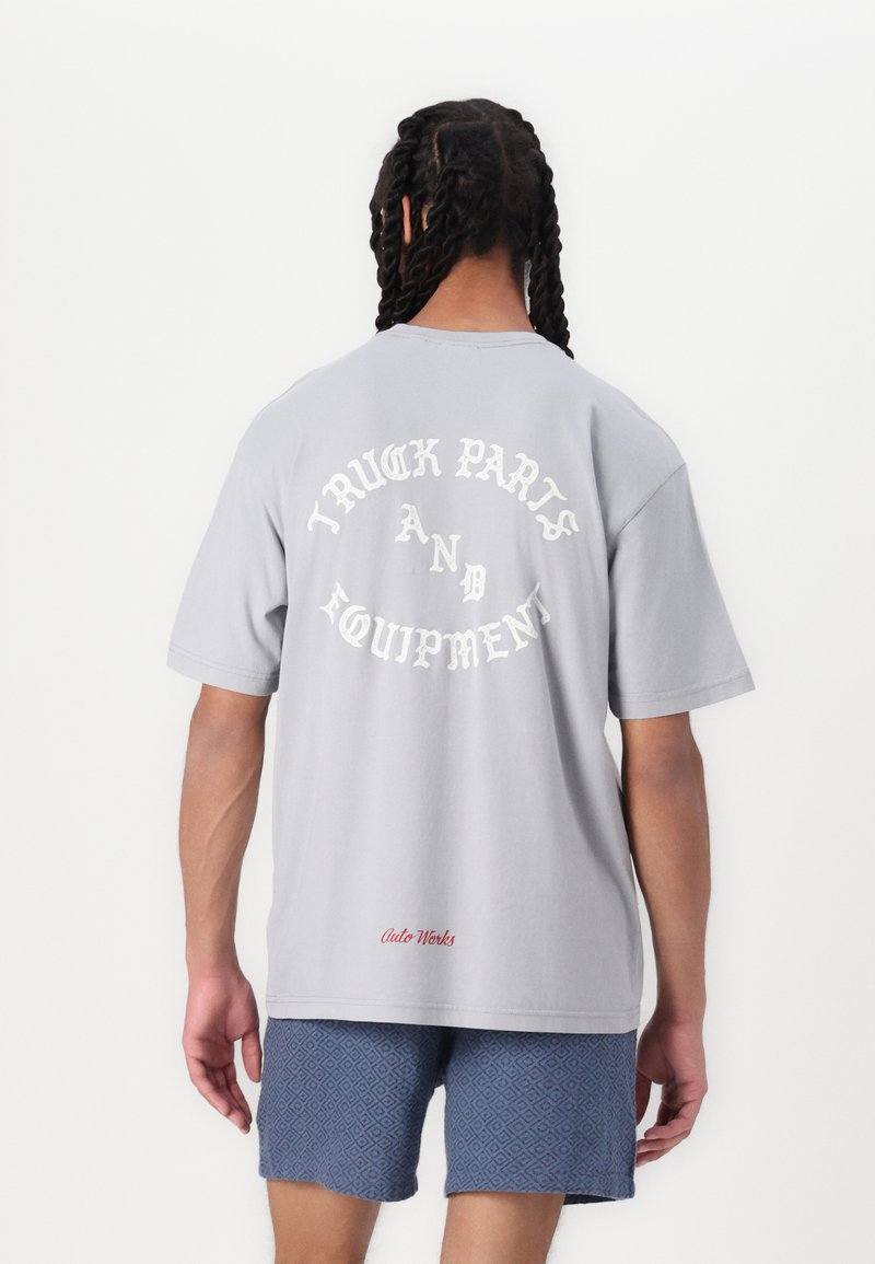 Szary bawełnowy t-shirt z krótkim rękawem, z białym napisem "CZĘŚCI I WYPOSAŻENIE CIĄGNIKÓW" w zakrzywionym wzorze na plecach. Czerwony detal "Auto Works".