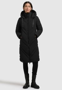 Kvinde iført en lang sort quiltet jakke med hætte, lynet op, hænderne i lommen, sammen med sorte leggings og sorte ankelstøvler.