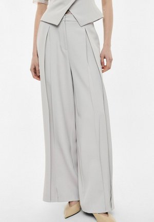 Pantaloni grigio chiaro a gamba larga con pieghe frontali, indossati con scarpe beige a punta e un gilet grigio chiaro abbinato.