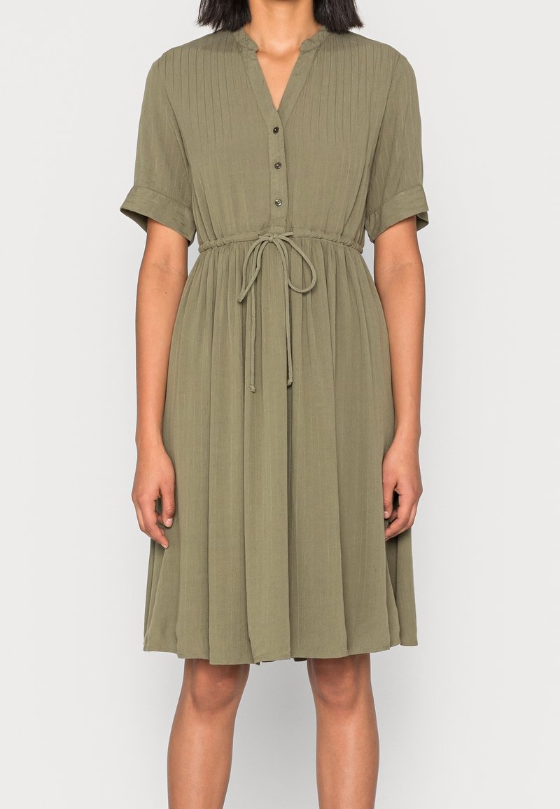 Femme portant une robe verte olive longueur genou avec manches courtes boutonnées et ceinture à cordon à la taille.