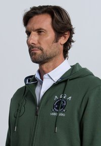 Sudadera con capucha verde con cremallera y camisa blanca con cuello debajo. Presenta un logo bordado y detalles en la cremallera, hecha de tela suave y texturizada.