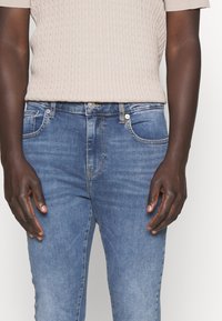 Jeans de mezclilla de color azul claro con un diseño clásico de cinco bolsillos, que cuentan con un cierre de botón y detalles de costura sutiles. Parte superior beige acanalada encima.