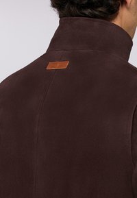 Giacca in suede marrone con colletto alto e una piccola etichetta in pelle sul retro. Texture liscia con dettagli di cucitura minimi.