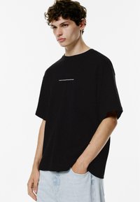 Sort bomulds T-shirt med en afslappet pasform, rund halsudskæring og korte ærmer. Har hvid tekst på forsiden. Bæres med lyseblå denimshorts.