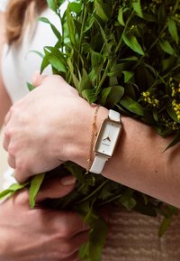 Montre à bracelet blanc avec un boîtier rectangulaire en or et un cadran blanc, accompagnée d'un délicat bracelet en or, maintenue parmi des feuillages verts.
