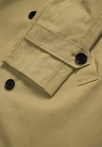 Khaki ballonkabát részlet, hajtott mandzsettával, fekete gombokkal és éles varrással, sima, texturált anyagot bemutatva.