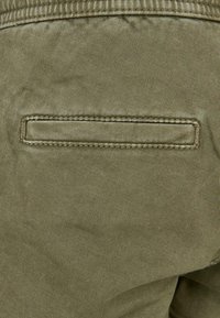 Tissu vert olive avec une texture côtelée verticale, présentant une poche cachée étroite et rectangulaire à l'arrière, cousue avec un fil assorti.