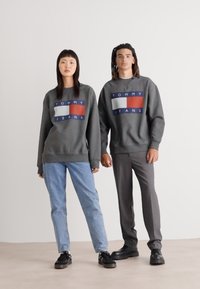 Šedé mikiny s logom "TOMMY JEANS", s červenými, bielymi a modrými akcentmi. V páre so svetlo modrými džínsami a tmavými topánkami.