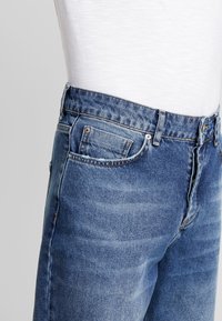 Blue denim jeans with a mid-rise waist, five-pocket design, and contrasting stitching. The fabric features a textured finish and slight fading.