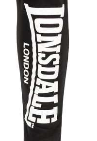 Zwarte sportbroek met grote witte tekst die "Lonsdale London" verticaal langs de zijkant leest, met een gladde katoenen textuur.