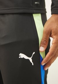 Pantalones deportivos negros con paneles laterales verdes y azules, textura suave y un logo blanco de Puma en la parte inferior izquierda.