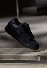 Schwarze Sneakers mit glattem Lederobermaterial, die ein Sternlogo an der Seite, runde Schnürsenkel und eine flache Gummisohle aufweisen, aus einem Winkel präsentiert.