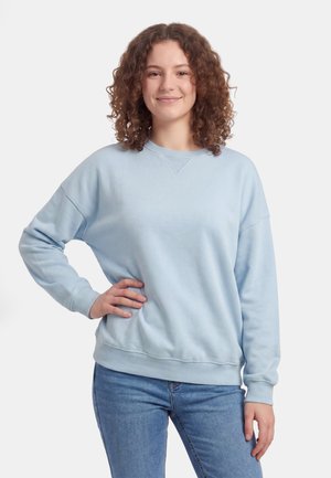 Młoda kobieta o kręconych włosach, ubrana w jasnoniebieską bluzę i niebieskie dżinsy, stoi z jedną ręką na biodrze, uśmiechając się.