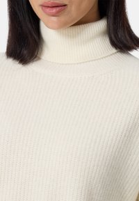Pull à col roulé blanc côtelé avec une texture douce et une silhouette ajustée, montrant le détail du cou et une partie du col.