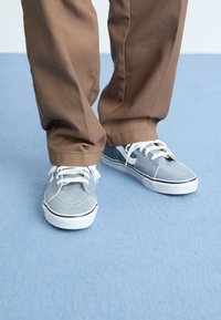 Grijze canvas sneakers met witte veters en zwarte accenten, gedragen met bruine pantalon. Focus op de ronde neus van de schoen en het gestructureerde oppervlak.