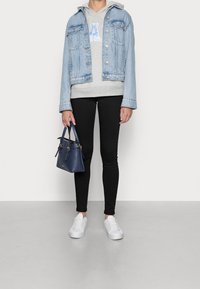 Chaqueta de mezclilla azul claro, sudadera con capucha gris con diseño de "A", jeans ajustados negros, zapatillas blancas, sujetando un pequeño bolso azul marino con detalles dorados.