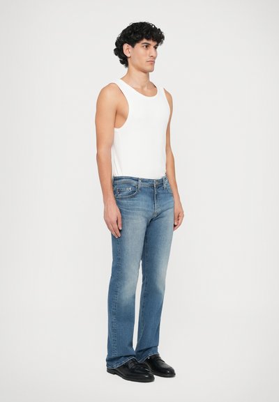 Weißes Tanktop, blaue, hell gewaschene Denim-Jeans, gerade Passform, mit Fronttaschen, dunkles Schuhwerk, Model steht seitlich vor einem hellen Hintergrund.