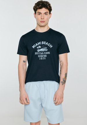 Młody mężczyzna z kręconymi włosami, ubrany w czarną koszulkę surfingową Miami Beach i jasnoniebieskie krótkie spodenki ze sznurkiem, stojący na jednolitym tle.