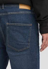 Temno modri denim kavbojke z zadnjim žepom, kontrastnim šivom in rjavim usnjenim etiketom. Tkanina prikazuje rahlo teksturo.