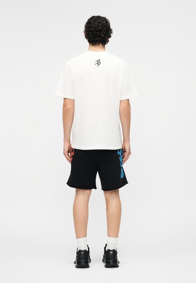 Άνδρας με σγουρά μαλλιά στέκεται με την πλάτη προς το μέρος μας, φοράει λευκό μπλουζάκι με μαύρο λογότυπο στο πάνω μέρος της πλάτης και μαύρο σορτς με κόκκινα και μπλε φλόγινα σχέδια.