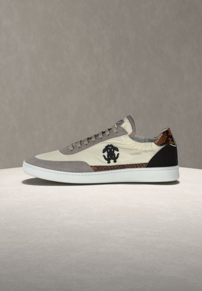 Χαμηλό sneaker με λευκή σόλα, επάνω μέρος από κρεμ ύφασμα, γκρι σουέτ λεπτομέρειες, μαύρο λογότυπο στο πλάι και καφέ σχέδιο δέρματος φιδιού κοντά στη φτέρνα και την περιοχή των κορδονιών.