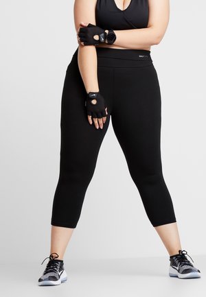 Femme en tenue sportive noire, portant des gants sans doigts et une montre connectée, debout jambes écartées contre un fond uni.