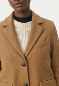 Brown wool-blend coat with a shawl collar, three buttons, and a single chest pocket. Worn over a cream cable-knit sweater with a black neck.