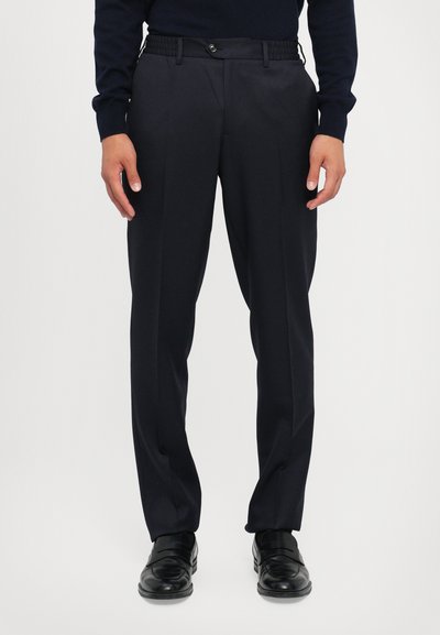 Pantaloni da cerimonia blu navy con una superficie liscia, dotati di una vita aderente, frontale piatto e pieghe nette. Indossati con scarpe di pelle nere.