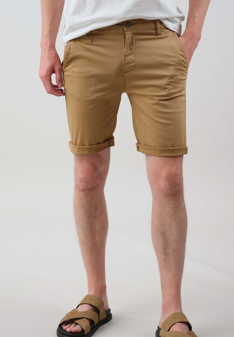 Shorts en khaki en mélange de coton avec des revers, dotés de poches avant et arrière, associés à des sandales à glissière marron.