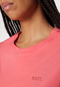 Gros plan d'une personne portant un T-shirt rose corail avec des cheveux tressés visibles et le mot « BOSS » brodé sur la poitrine.